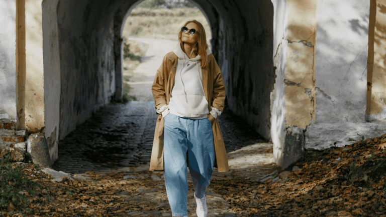 Frau mit Sonnenbrille, braunem Mantel, Hoodie und Jeans schlendert durch den Herbst