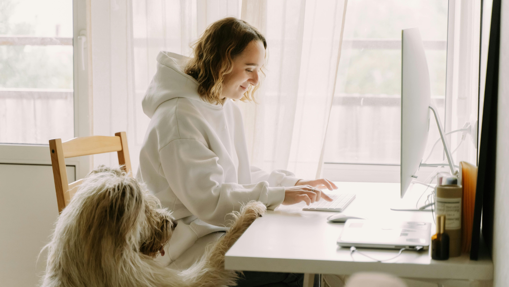 Frau mit weißem Hoodie sitzt am Computer, Hund schaut am Schreibtisch hoch