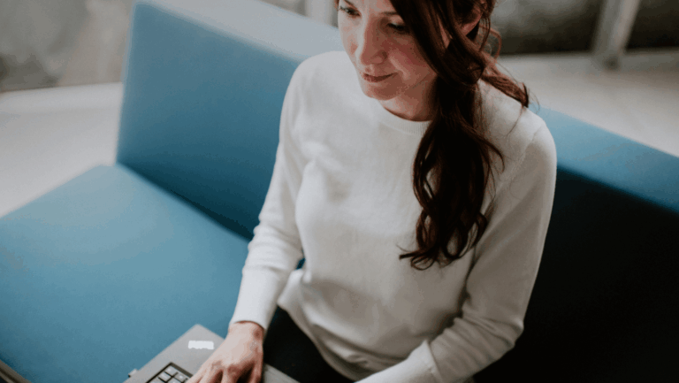 Frau mit langen dunklen Haaren sitzt am Laptop auf einem blauen Sofa