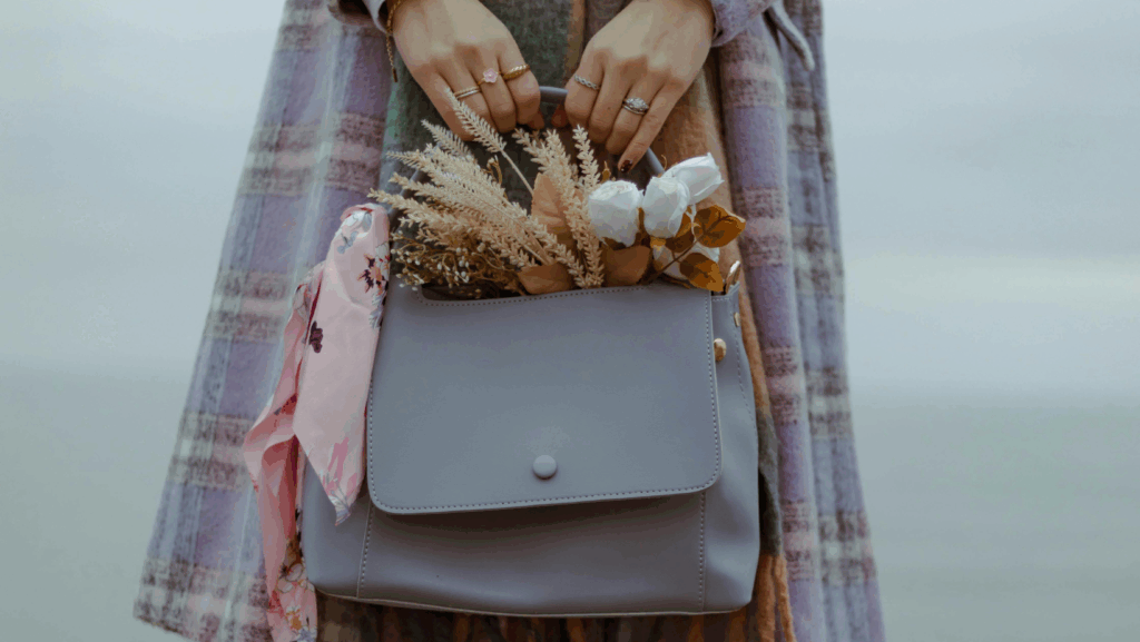 Frau hält graue Handtasche in den Händen, aus der Blumen, Weizen und ein Tuch herausgucken