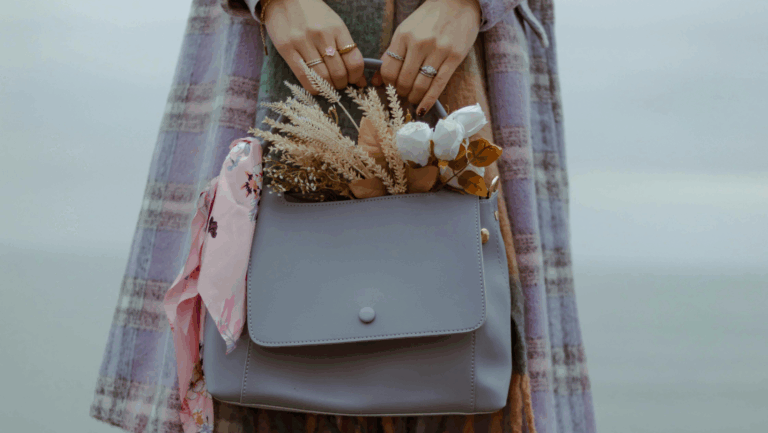 Frau hält graue Handtasche in den Händen, aus der Blumen, Weizen und ein Tuch herausgucken