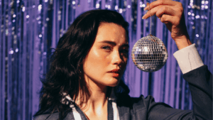 Junge dunkelhaarige Frau steht mit einer kleinen Diskokugel neben ihrem Kopf vor einem lilanen Glitzer-Vorhang