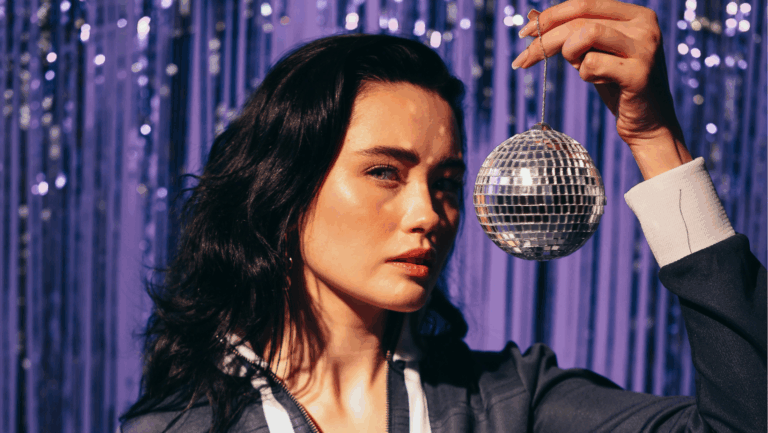 Junge dunkelhaarige Frau steht mit einer kleinen Diskokugel neben ihrem Kopf vor einem lilanen Glitzer-Vorhang