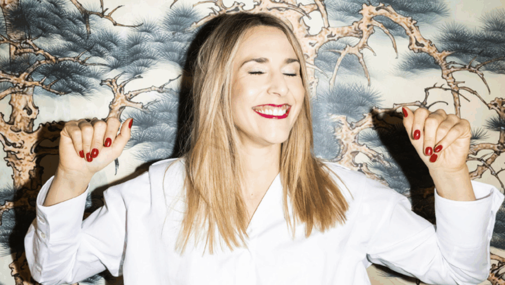 Aimie Carstensen strahlt mit geschlossenen Augen in einer weißen Bluse mit rotem Lippenstift