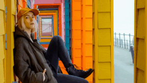 Junge, blonde Frau sitzt mit einem Cap vor einer bunten Kulisse