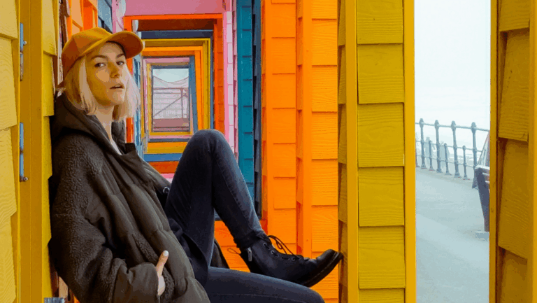 Junge, blonde Frau sitzt mit einem Cap vor einer bunten Kulisse