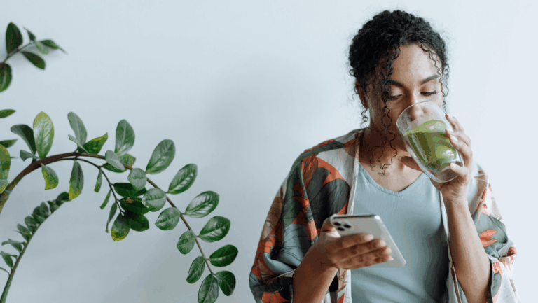 Junge Frau ist zur Hälfte zu sehen, sie guckt auf ihre Handy und trinkt dabei einen grünen Smoothie