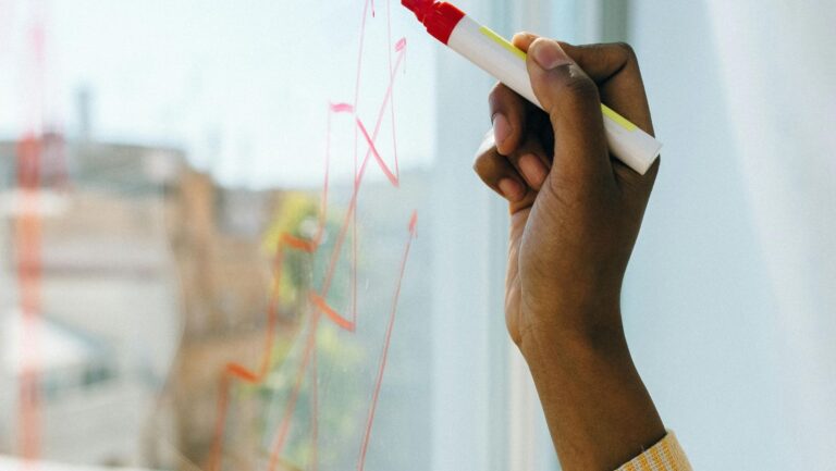 Eine Hand zeichnet mit einem roten Stift einen Aktienkurs auf die Fensterscheibe