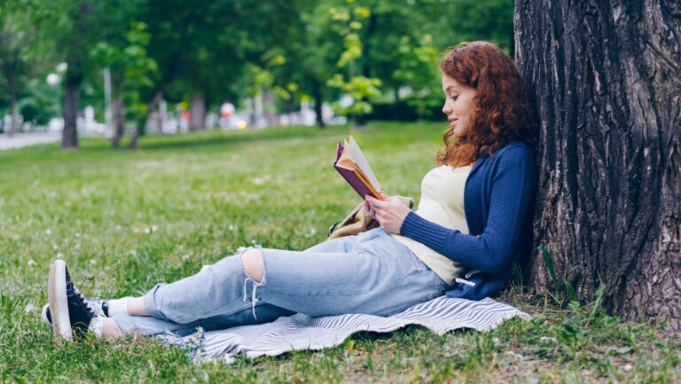 Eine junge Frau sitzt angelehnt an einen Baum im Park und liest ein Buch