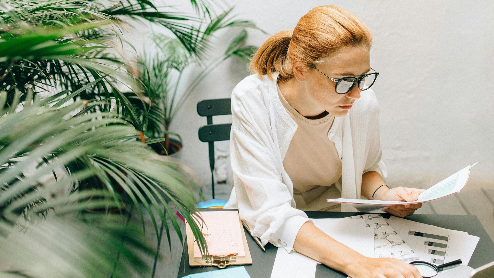 Frau mit Brille sitzt am Schreibtisch und guckt auf einen Zettel