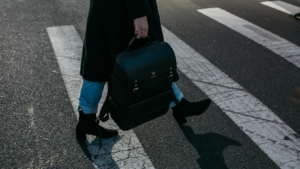 Eine Frau, bekleidet mit Jeans, schwarzen Absatzstiefeln und schwarzem Mantel läuft mit einer Aktentasche über einen Zebrastreifen. Ihren Kopf sieht man nicht, er ist im Bild abgeschnitten.