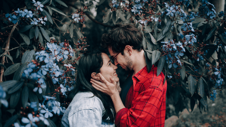 Ein junges Paar: Er nimmt ihren Kopf in seine Hände, beide sehen sich an und stehen vor einem grünen Blätterwerk