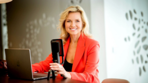 Carola Ferstl: Die Finanzjournalistin sitzt in einem orangenen Blazer vor einem Mikrofon und lächelt in die Kamera.