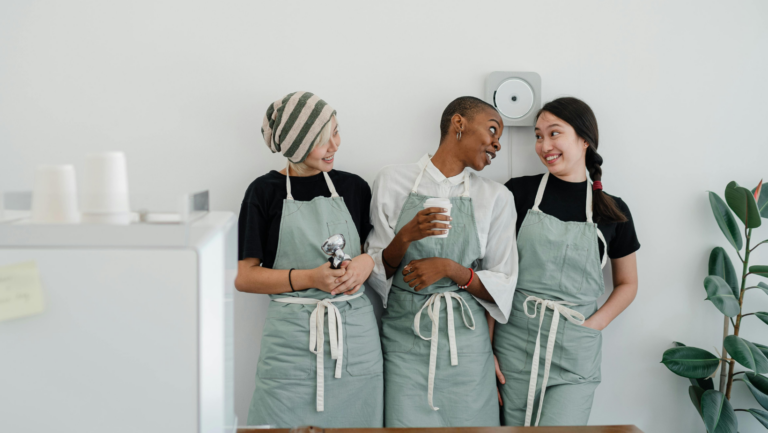 Drei junge Frauen in hellgrüner Schürze stehen in einer Küche eng nebeneinander, zwei von ihnen gucken sich an, die dritte sieht in Richtung der beiden anderen.