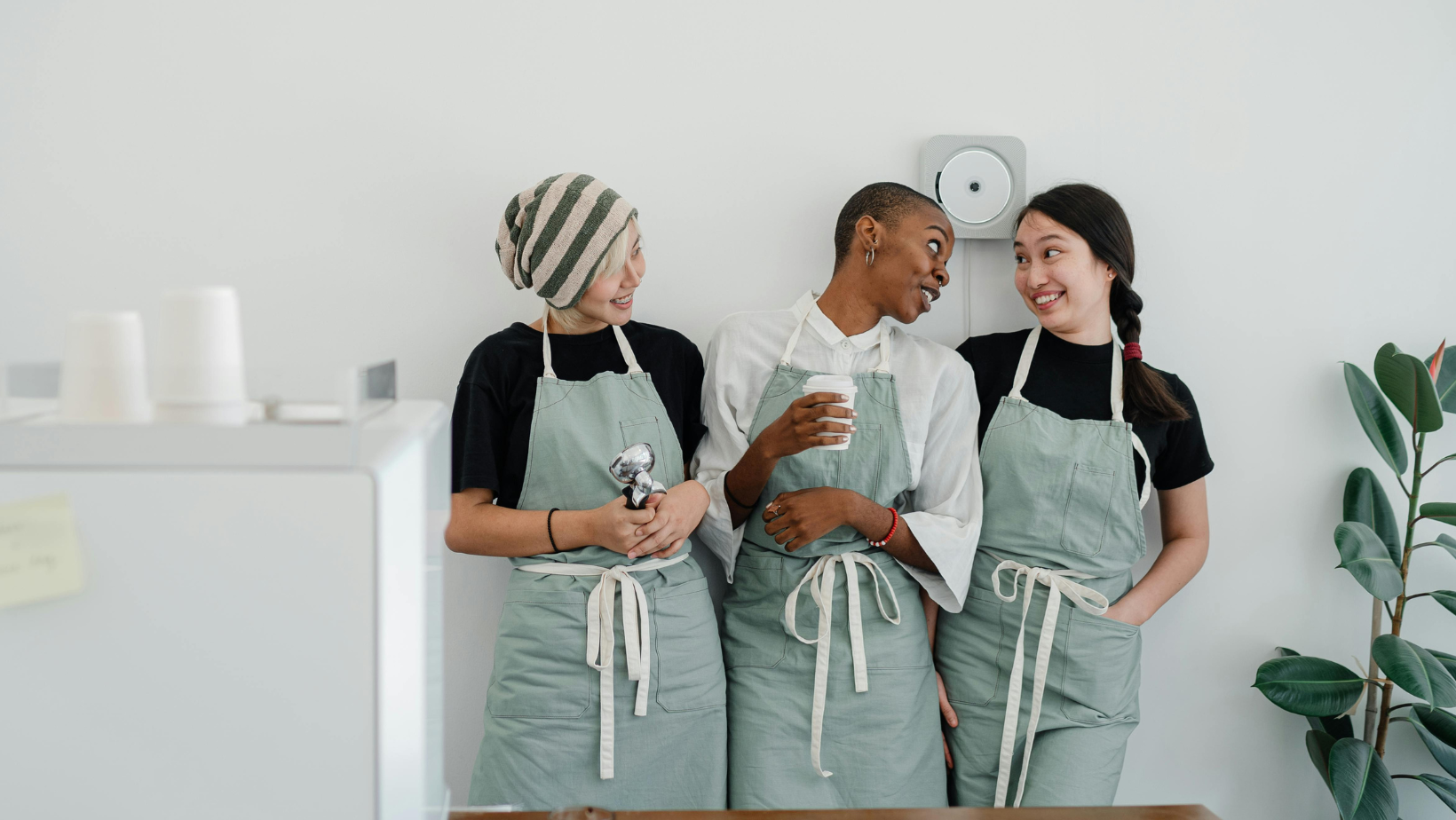 Drei junge Frauen in hellgrüner Schürze stehen in einer Küche eng nebeneinander, zwei von ihnen gucken sich an, die dritte sieht in Richtung der beiden anderen.