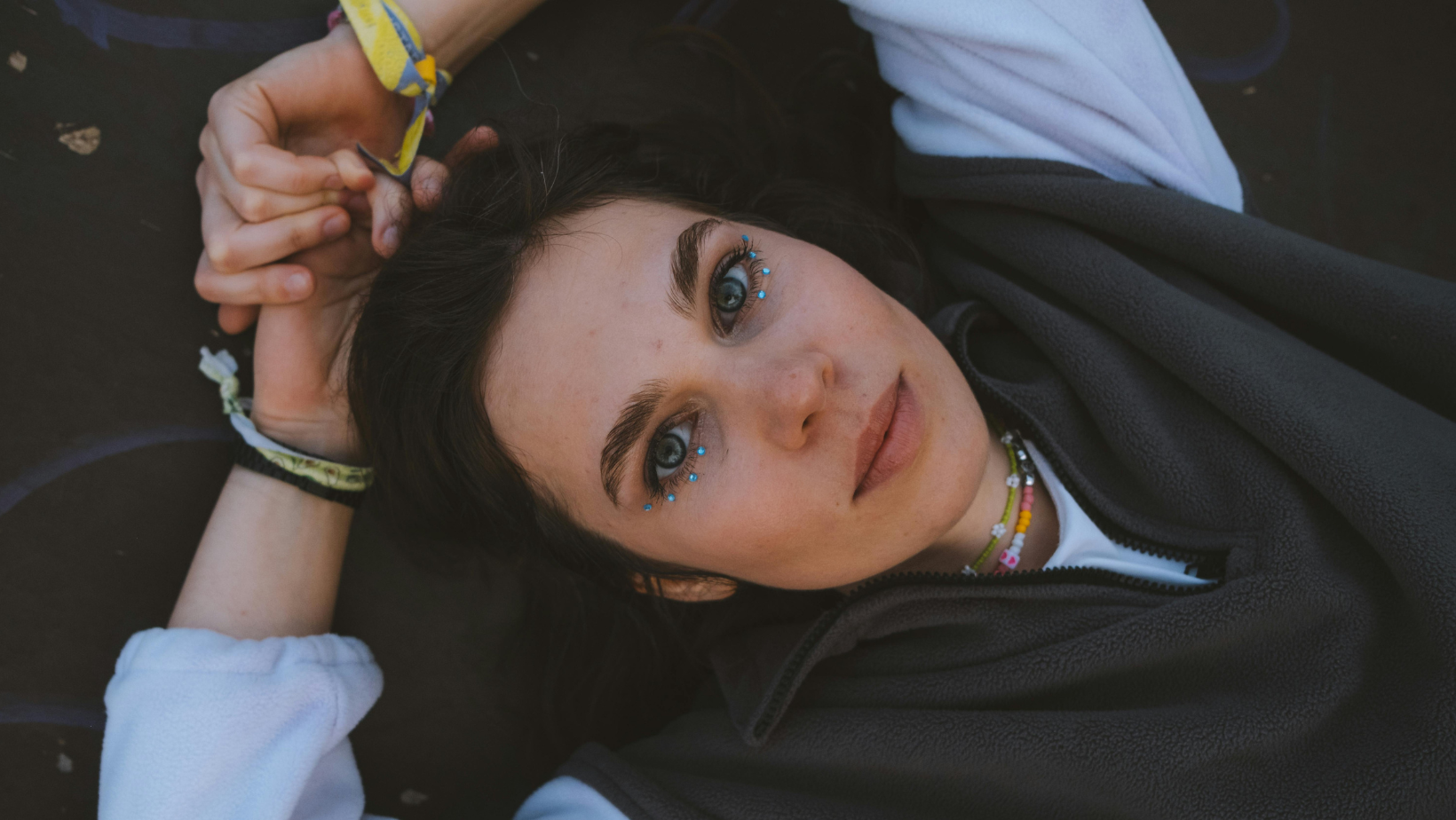 Junge Frau liegt und schaut in die Kamera. Sie hat dunkle Haare und unter den Augen blaue Glitzersteine.