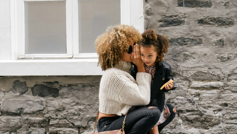 POC-Frau mit Sonnenbrille hat ihre etwa 7-jährige Tochter auf dem Arm und flüstert ihr mit vorgehaltener Hand etwas ins Ohr