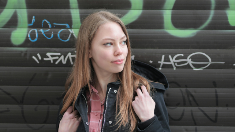 Junge Frau schaut zur Seite, hinter ihr sieht man eine Außenjalousie, die mit Graffity besprüht wurde