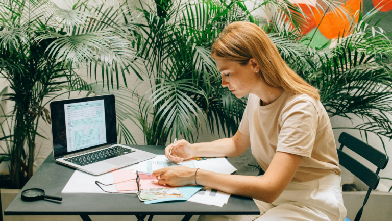 Frau sitzt am Schreibtisch und liest Abrechnungen, im Hintergrund ein aufgeklappter Laptop