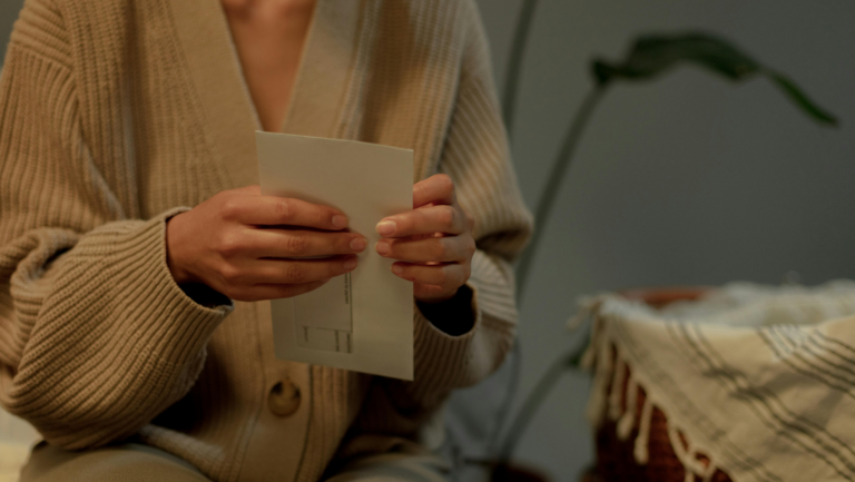 Eine Frau hält einen Brief in der Hand. Zu sehen sind aber nur ihre Hände und ihr Oberkörper in einer beigen Strickjacke