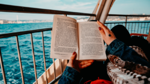 Aufgeschlagenes Buch wird von zwei Händen gehalten, im Hintergrund ein Schiffsgeländer und blaues Meer
