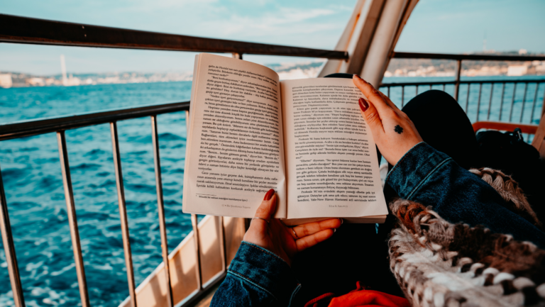 Aufgeschlagenes Buch wird von zwei Händen gehalten, im Hintergrund ein Schiffsgeländer und blaues Meer