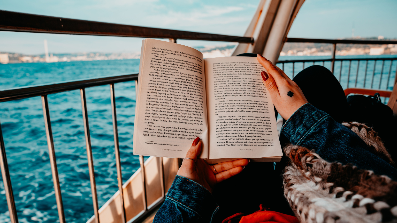 Aufgeschlagenes Buch wird von zwei Händen gehalten, im Hintergrund ein Schiffsgeländer und blaues Meer