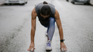 Frau hockt am Boden in der Styrtposition für einen Sprint. Sie trägt graue Sportkleidung, man sieht den Kopf nur von oben, kein Gesicht.