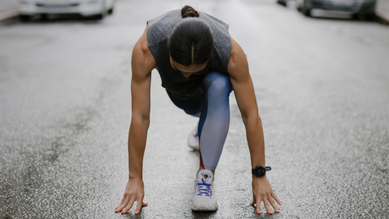 Frau hockt am Boden in der Styrtposition für einen Sprint. Sie trägt graue Sportkleidung, man sieht den Kopf nur von oben, kein Gesicht.