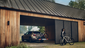 Ein Holzhaus mit Zinkdach und einer Garage, in der ein Auto mit einem Thule Fahrradträger steht. Vor dem Haus sieht man eine Frau mit ihrem Fahrrad.