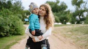 Eine junge Frau mit langen Locken hält ein etwa zweijähriges Kind mit Mütze auf dem Arm