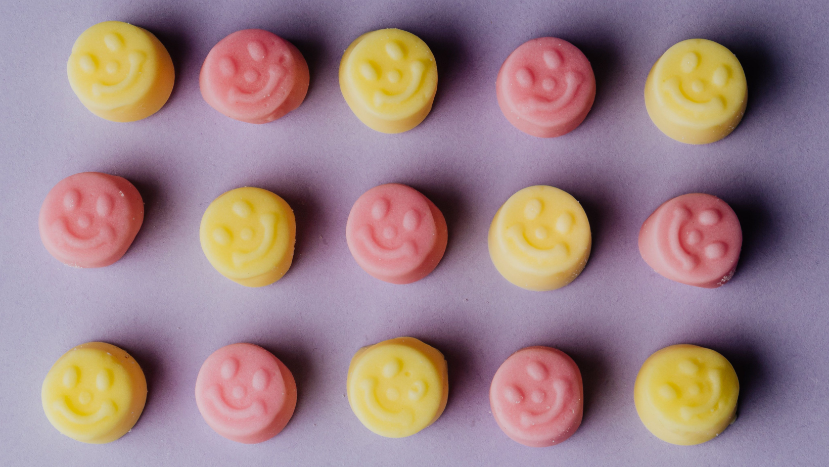 Auf einem violetten Hintergrund liegen abwechselnd pinke und gelbe runde Smileys.