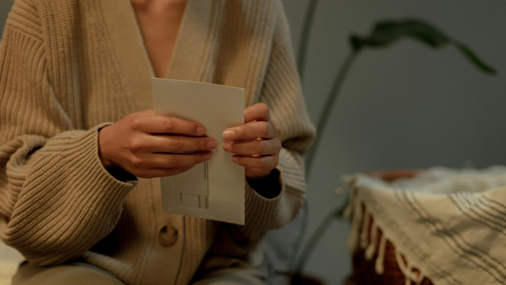 Eine Frau hält einen Brief in der Hand. Zu sehen sind aber nur ihre Hände und ihr Oberkörper in einer beigen Strickjacke